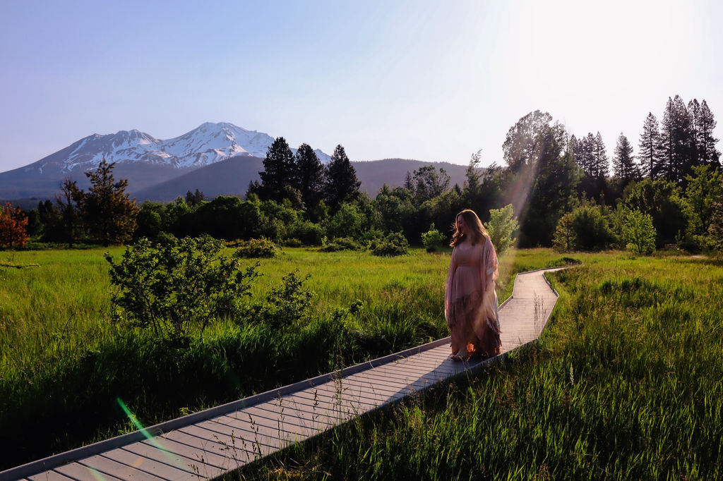 A woman walking a wooden boardwalk through a green meadow below Mount Shasta, California